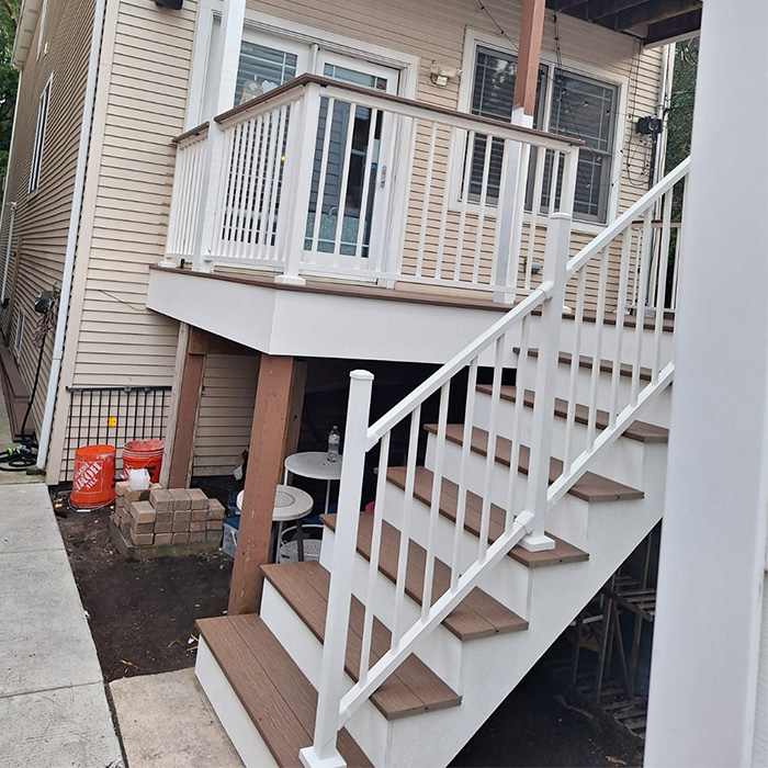 Patio elevado con barandilla blanca y escaleras, adosado a una casa beige. Debajo, se ven una mesa pequeña y ladrillos. Ambiente residencial urbano.