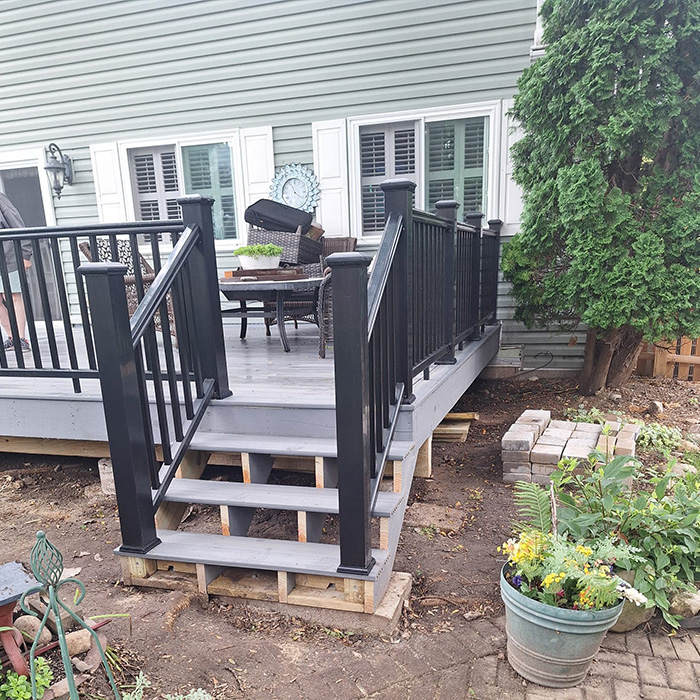 Una terraza de madera gris con barandillas negras da paso a una casa con revestimiento verde. Muebles de jardín, plantas en macetas y un árbol bien cuidado crean un ambiente acogedor.