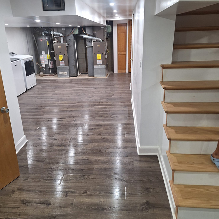 Un sótano con suelo de madera oscura, con escaleras de madera a la derecha. Tres hornos metálicos bordean la pared del fondo, junto a una lavadora y una secadora.