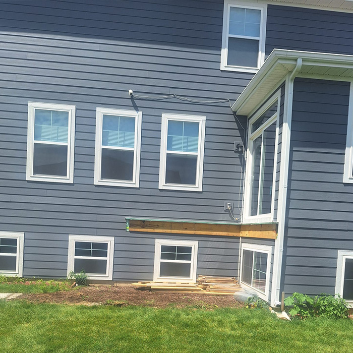 Vista exterior de una casa gris con múltiples ventanas, un alero de madera y un patio ajardinado con algunos materiales de construcción.