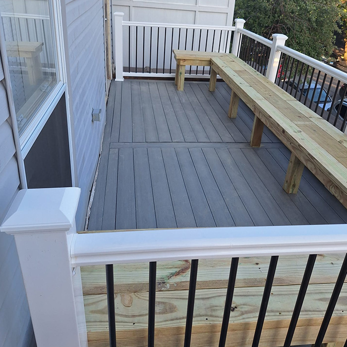 Una terraza de madera con tablones gris claro, barandilla blanca y balaustres negros. Un sencillo banco de madera recorre el lado derecho. El ambiente es tranquilo y ordenado.