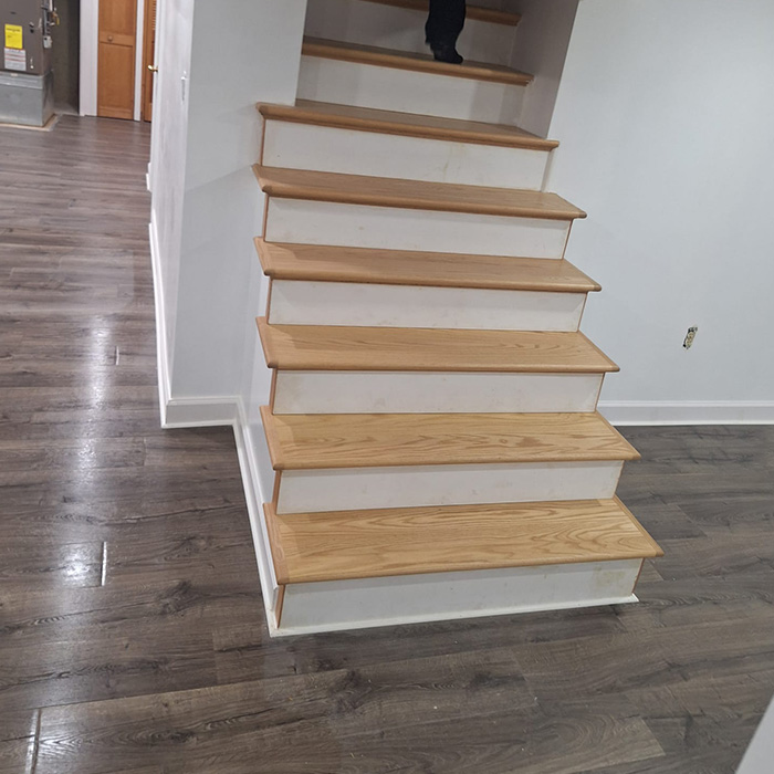 Escalera de madera con peldaños marrón claro y contrahuellas blancas, en una habitación con suelo de madera oscura y paredes blancas lisas. Una persona está de pie en lo alto.