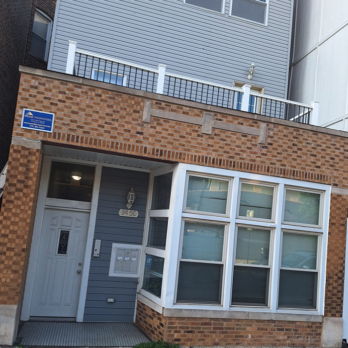 Edificio de dos plantas de ladrillo y revestimiento, con amplios ventanales. La entrada cuenta con una puerta blanca y buzones. Un cartel de seguridad visible transmite un ambiente seguro.