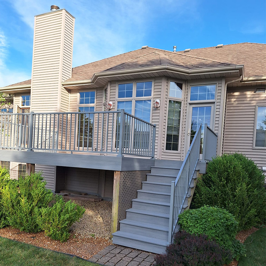 Una casa moderna con patio y escaleras que bajan a un jardín ajardinado, con chimenea y grandes ventanales. Cielo azul brillante.