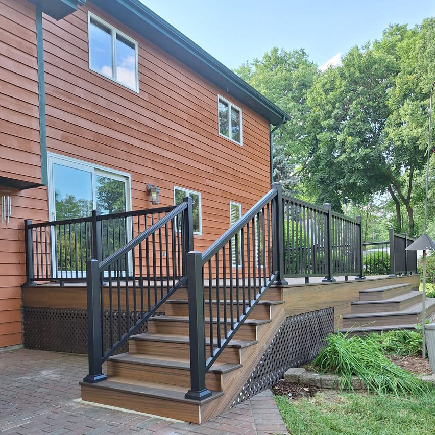 Una terraza de madera con barandillas y escaleras negras, adosada a una casa con revestimiento marrón, rodeada de exuberante vegetación y árboles.