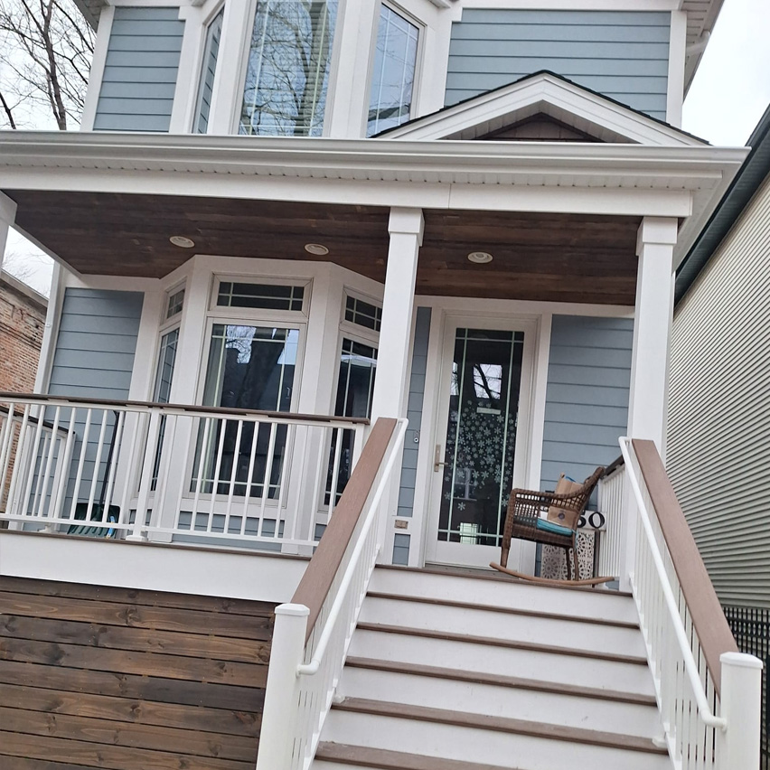 Casa azul moderna con porche de madera, barandillas blancas y escalones que conducen a una puerta de cristal. Hay una mecedora en el porche.